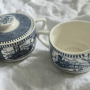 Op de afbeelding: Een keramische suikerpot met deksel en een bijpassende melkkan, beide met een witte basis en blauwe transferware-patronen van een stoomlocomotief en een stoomboot, afgewerkt met decoratieve geschulpte randen.