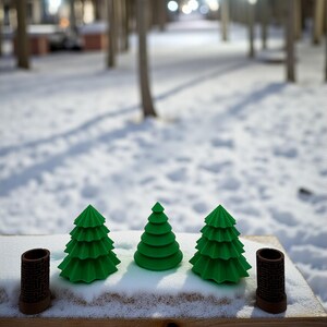 May include: Three green Christmas tree figurines and two brown cylindrical objects with a maze pattern are arranged on a wooden surface covered in snow. The background shows a snowy path and trees.
