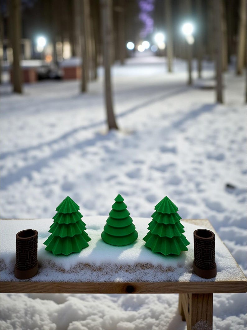 May include: Three green Christmas tree figurines and two brown cylindrical objects with a maze pattern are arranged on a wooden surface covered in snow. The background shows a snowy path and trees.