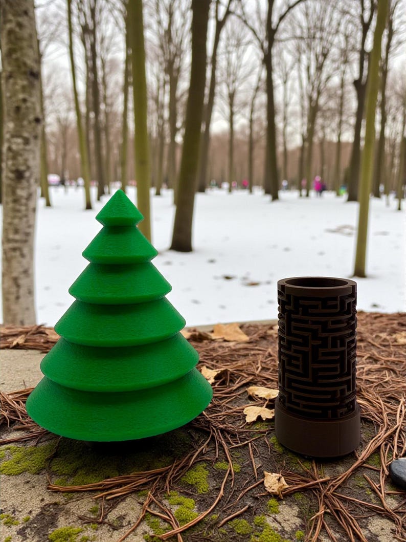 May include: A green, tiered Christmas tree and a brown cylindrical object with a maze design sit on a mossy surface. The background shows a snowy park with bare trees.