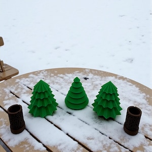 May include: Three green, stylized Christmas tree decorations and two brown cylindrical holders sit on a snow-covered wooden table. The trees have a layered design. The holders have a geometric pattern.