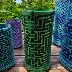 May include: A collection of cylindrical planters featuring intricate, raised maze patterns on the exterior. The planters are finished in various gradient colors, including metallic green, purple, and blue, with a diamond-textured base design.