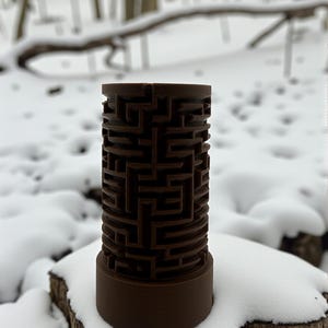 May include: A brown cylindrical puzzle with a maze design. The maze is carved into the exterior of the cylinder. The puzzle is sitting on a snow-covered tree stump, with a snowy forest in the background.