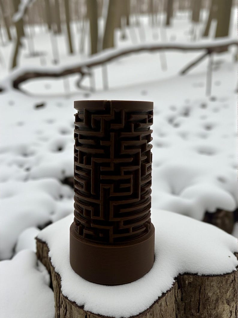 May include: A brown cylindrical puzzle with a maze design. The maze is carved into the exterior of the cylinder. The puzzle is sitting on a snow-covered tree stump, with a snowy forest in the background.
