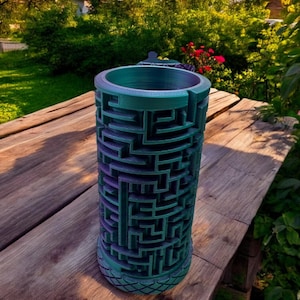 May include: A cylindrical container featuring a complex, three-dimensional maze pattern on its exterior surface. The object displays a gradient color shift from deep purple to teal, with a textured, diamond-patterned base and a smooth, circular rim.