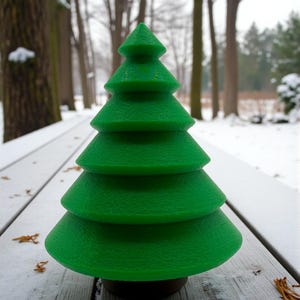 May include: A bright green, tiered Christmas tree decoration. The tree has a textured surface and a dark brown base. It is sitting on a weathered, white wooden surface with snow and fallen leaves in the background.