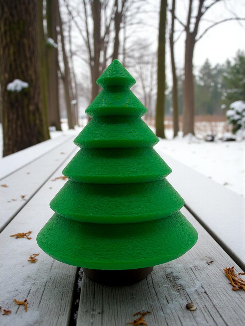 May include: A bright green, tiered Christmas tree decoration. The tree has a textured surface and a dark brown base. It is sitting on a weathered, white wooden surface with snow and fallen leaves in the background.