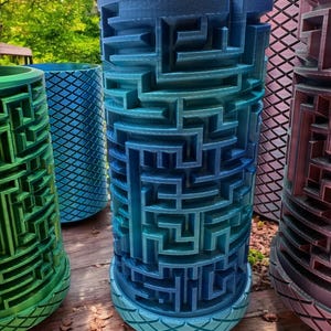 May include: A cylindrical 3D printed object featuring a complex, raised maze pattern on its exterior surface. The object displays a gradient of blue colors, transitioning from a lighter shade at the base to a deeper blue at the top, with a diamond-textured rim.