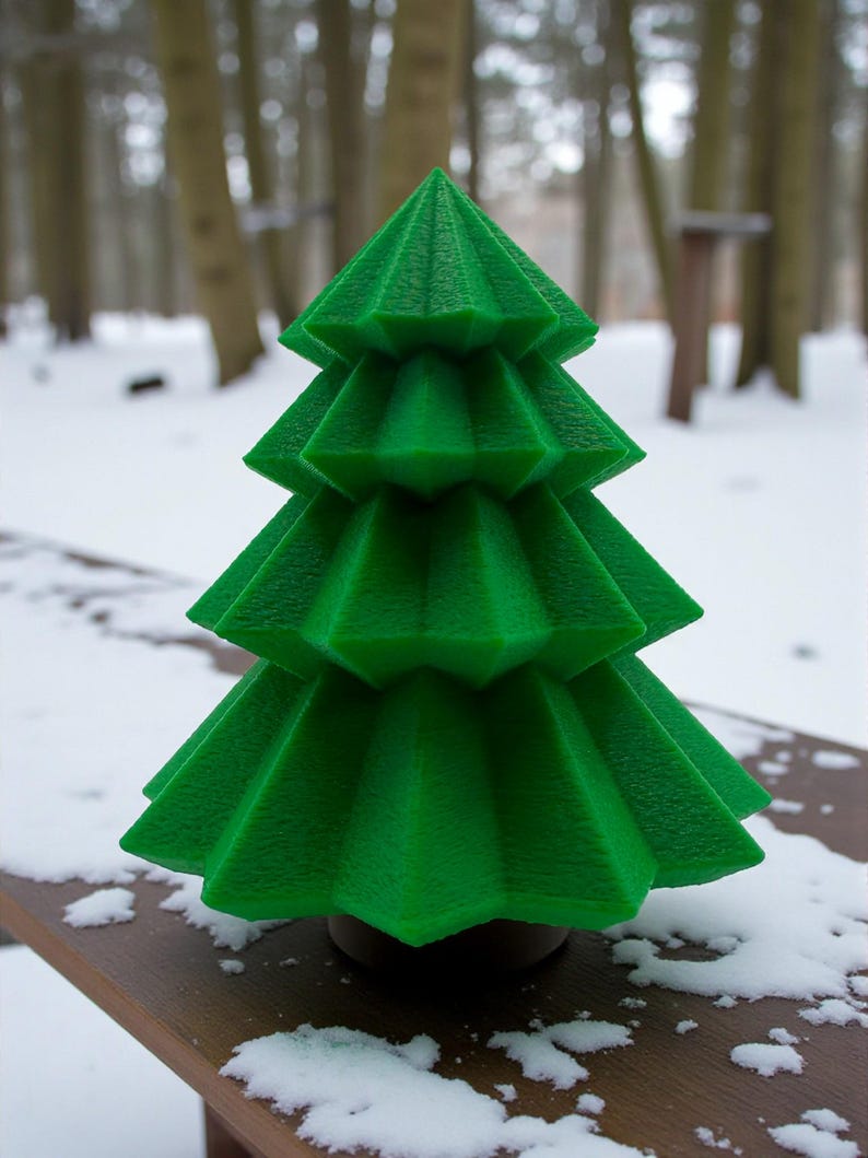 May include: A vibrant green, geometric Christmas tree decoration. The tree has a layered design with textured surfaces, set against a snowy outdoor backdrop. The tree is approximately 6 inches tall.