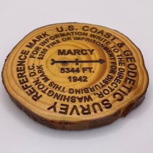 Puede incluir: Un disco de madera con el texto "U.S. Coast & Geodetic Survey" y "Reference Mark for Information. $250 Fine or Imprisonment for Disturbing This Mark. Write to the Director, Washington D.C." El disco tambi&eacute;n tiene el texto "Marcy 5344 Ft. 1942" y una flecha que apunta hacia la derecha.