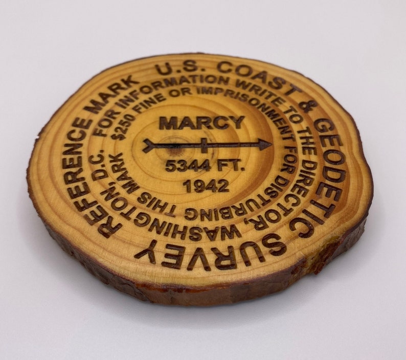Puede incluir: Un disco de madera con el texto "U.S. Coast & Geodetic Survey" y "Reference Mark for Information. $250 Fine or Imprisonment for Disturbing This Mark. Write to the Director, Washington D.C." El disco tambi&eacute;n tiene el texto "Marcy 5344 Ft. 1942" y una flecha que apunta hacia la derecha.