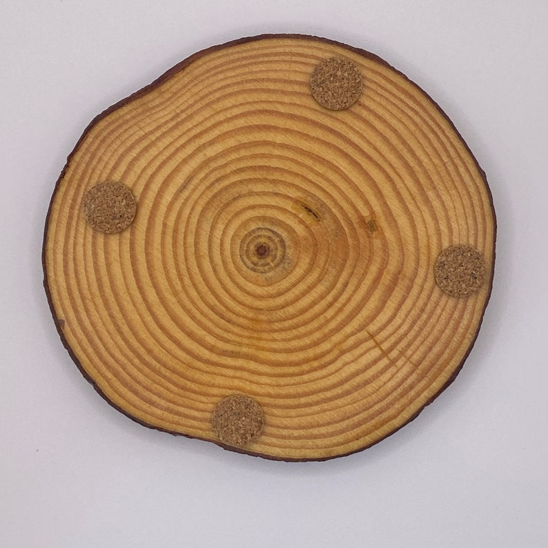 Puede incluir: Un posavasos redondo de madera con un acabado de grano de madera natural. El posavasos tiene cuatro almohadillas de fieltro marr&oacute;n en la parte inferior.