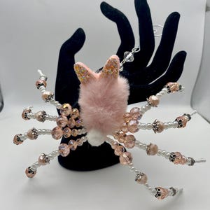 May include: A unique, handmade spider ornament with a pink bunny head and fluffy body. The spider's legs are made of white pearls, pink beads, and skull-shaped beads. The bunny ears are pink with glitter.