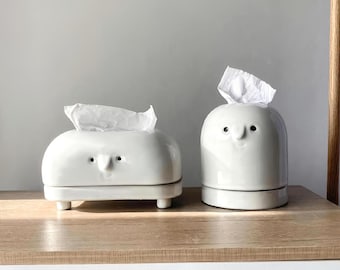 Puede incluir: Dos porta pañuelos de cerámica blanca con un diseño de cara de dibujos animados. Un porta pañuelos es de forma cuadrada con patas y el otro es de forma redonda.