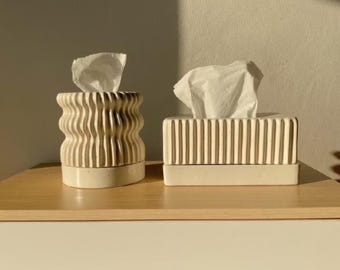Puede incluir: Dos porta pañuelos de cerámica blanca. Uno tiene un diseño ondulado y el otro tiene líneas verticales. Ambos están sobre una superficie de madera.