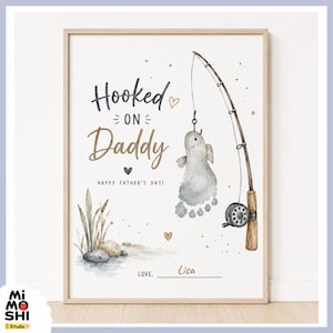 May include: A framed print with the text "Hooked on Daddy" and "Happy Father's Day!" features a watercolor illustration of a fishing rod with a fish-shaped footprint. The artwork includes a fishing reel, reeds, and rocks.