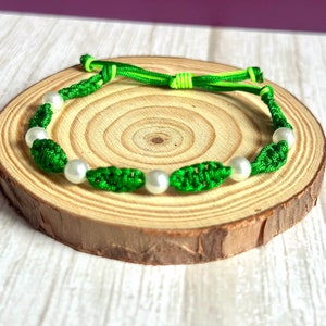 May include: A green and white bracelet with white pearl beads and green knotted cord. The bracelet is on a wooden surface.