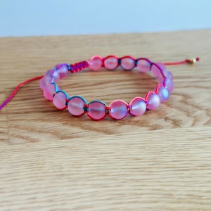 Puede incluir: Un brazalete de cuentas iridiscentes de color rosa con detalles de cuerda roja, azul y morada.