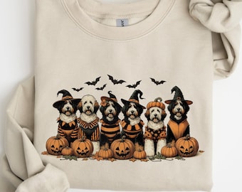 May include: Beige sweatshirt featuring a Halloween-themed graphic. The design includes six dogs dressed in costumes, wearing witch hats and pumpkin outfits, with pumpkins and bats. A cozy and festive apparel item.