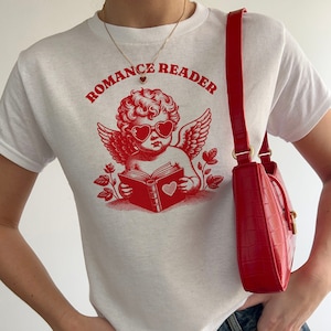 May include: White t-shirt with a red graphic of a cherub wearing heart-shaped sunglasses and reading a book. The text "Romance Reader" is printed above the cherub.