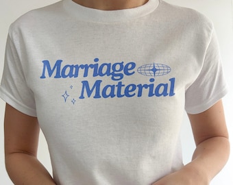 Puede incluir: Camiseta blanca con texto azul que dice "Marriage Material" con un icono de globo azul y dos estrellas azules.