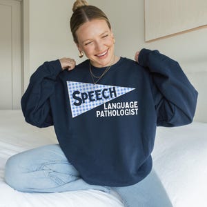 Könnte beinhalten: Marineblaues Sweatshirt mit dem weißen Schriftzug "SPEECH LANGUAGE PATHOLOGIST". Der Text befindet sich in einem weiß-blauen Karodreieck. Das langärmlige Sweatshirt wird mit einer blauen Jeans getragen.