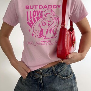 Puede incluir: Camiseta rosa con un estampado gr&aacute;fico de la cara de una mujer y el texto "But Daddy I Love Him" y "Broken Hearts Club 24/7 36".