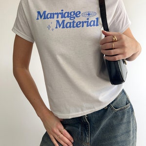 Puede incluir: Camiseta blanca con texto azul que dice "Marriage Material" y un gr&aacute;fico de globo terr&aacute;queo azul.