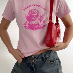 May include: Pink t-shirt with a graphic of a cherub wearing heart-shaped sunglasses and reading a book with a heart on the cover. The text "ROMANCE READER" is printed in a circle around the cherub.