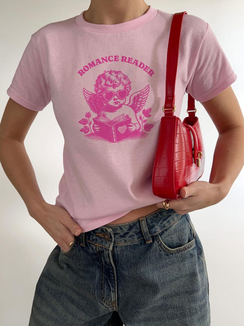 May include: Pink t-shirt with a graphic of a cherub wearing heart-shaped sunglasses and reading a book with a heart on the cover. The text "ROMANCE READER" is printed in a circle around the cherub.