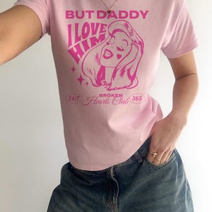 Puede incluir: Camiseta rosa con un gr&aacute;fico de una mujer con el texto "But Daddy I Love Him" y "Broken Hearts Club" impreso en ella.