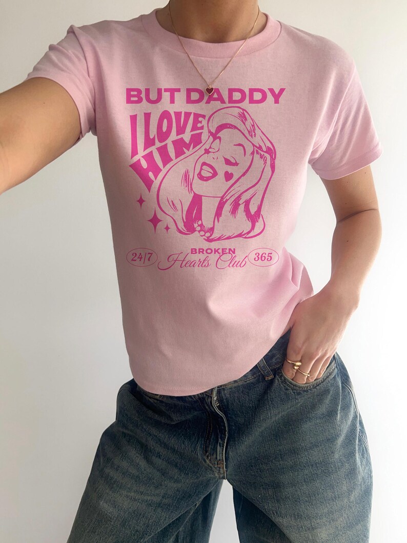 Puede incluir: Camiseta rosa con un gr&aacute;fico de una mujer con el texto "But Daddy I Love Him" y "Broken Hearts Club" impreso en ella.