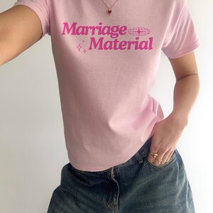 Puede incluir: Camiseta rosa con el texto "Marriage Material" en letras rosas con un peque&ntilde;o coraz&oacute;n rosa y estrellas impresas en la parte delantera.