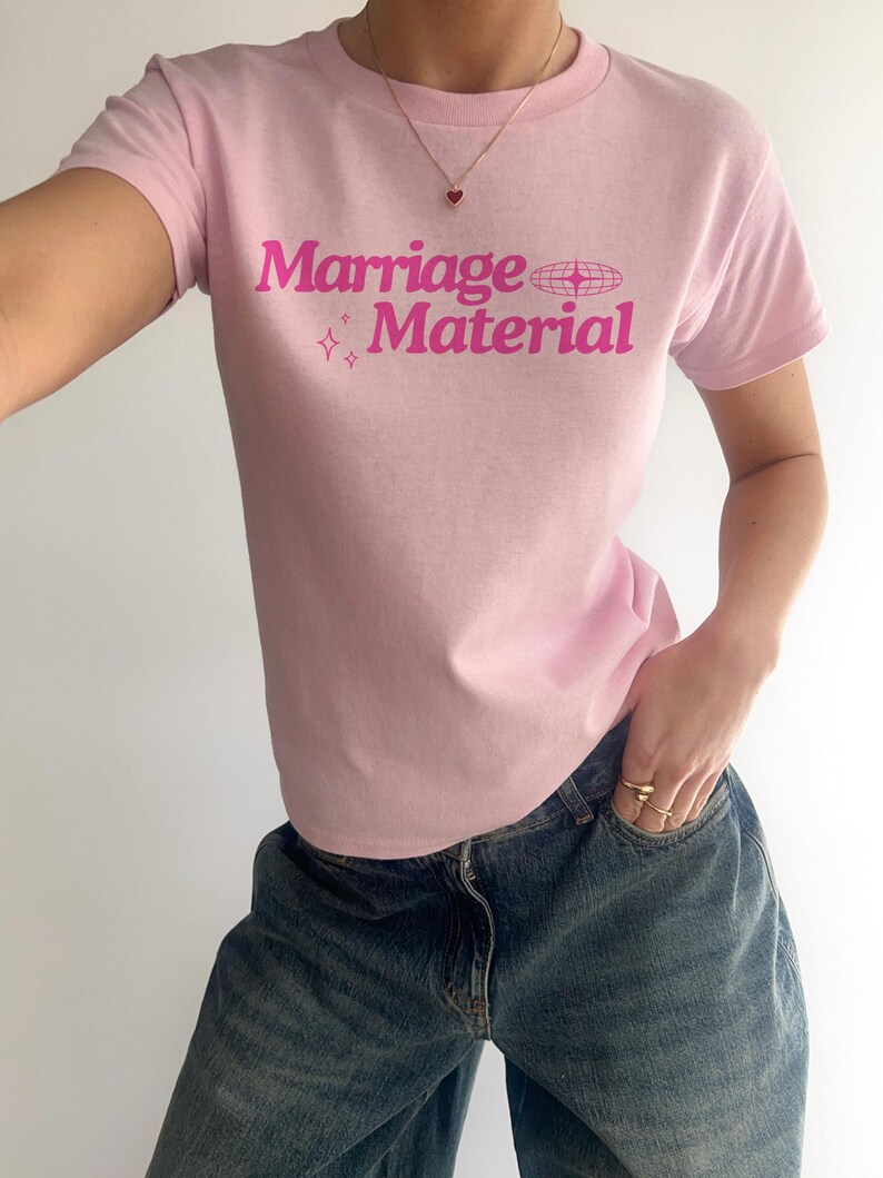 Puede incluir: Camiseta rosa con el texto "Marriage Material" en letras rosas con un peque&ntilde;o coraz&oacute;n rosa y estrellas impresas en la parte delantera.