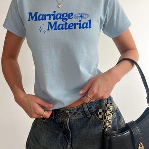 Puede incluir: Una camiseta azul claro con el texto "Marriage Material" en letras azules. La camiseta tiene un peque&ntilde;o gr&aacute;fico de globo terr&aacute;queo y dos estrellas.