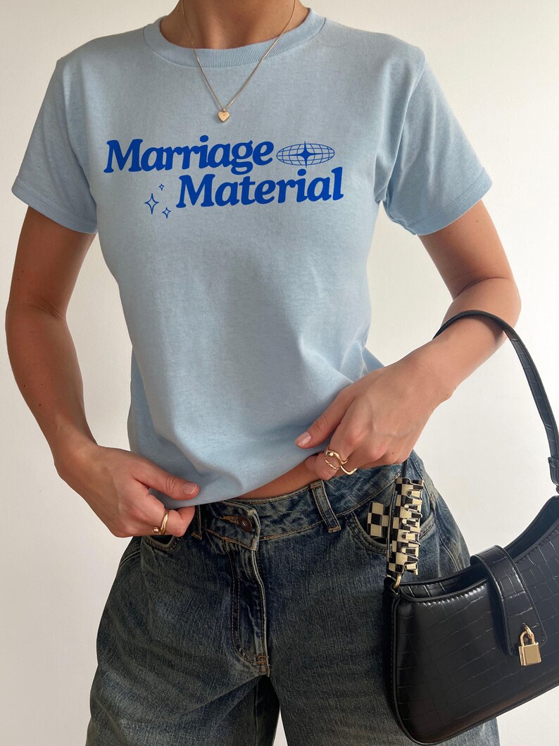 Puede incluir: Una camiseta azul claro con el texto "Marriage Material" en letras azules. La camiseta tiene un peque&ntilde;o gr&aacute;fico de globo terr&aacute;queo y dos estrellas.