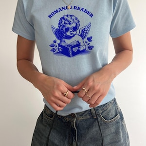 May include: A light blue t-shirt with a graphic of a cherub wearing sunglasses and reading a book. The text "ROMANCE READER" is printed in blue above the cherub.