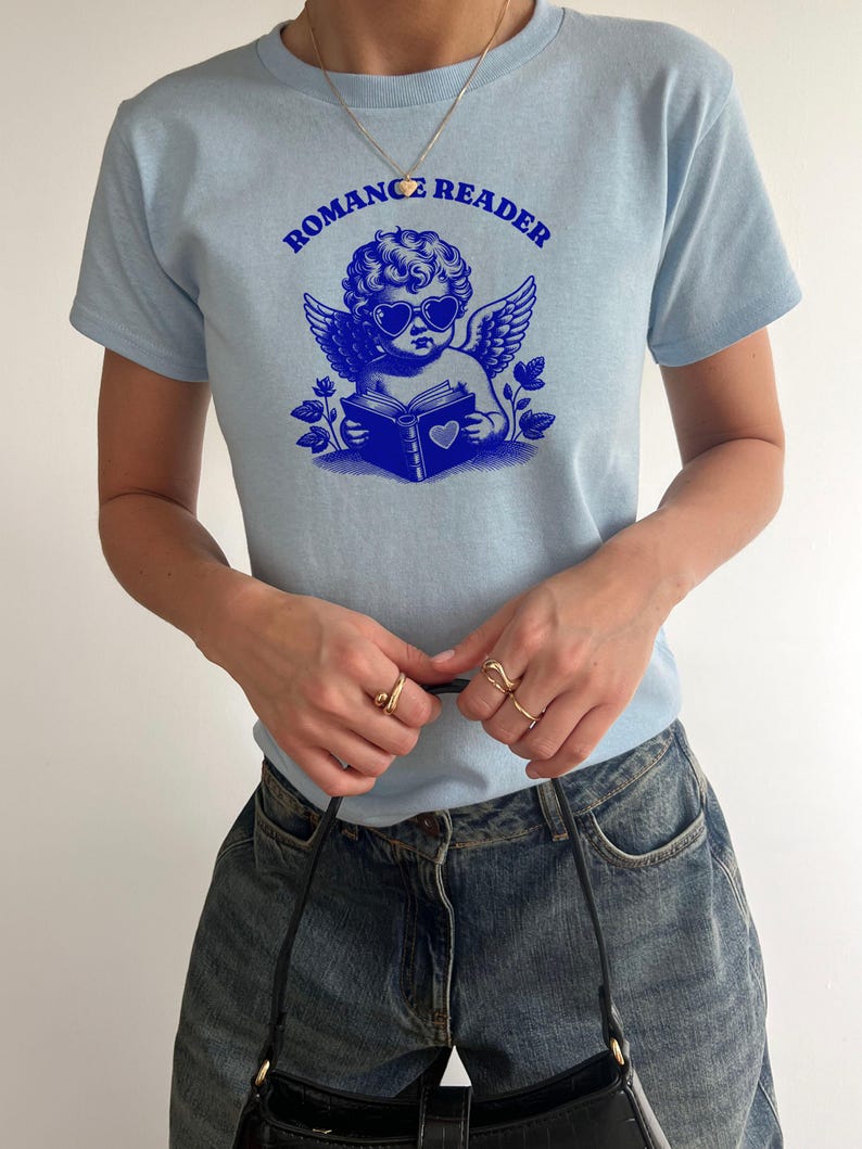 May include: A light blue t-shirt with a graphic of a cherub wearing sunglasses and reading a book. The text "ROMANCE READER" is printed in blue above the cherub.
