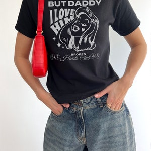 Puede incluir: Camiseta negra con un estampado gr&aacute;fico de la cara de una mujer y el texto "But Daddy I Love Him 24/7 Broken Hearts Club 365".