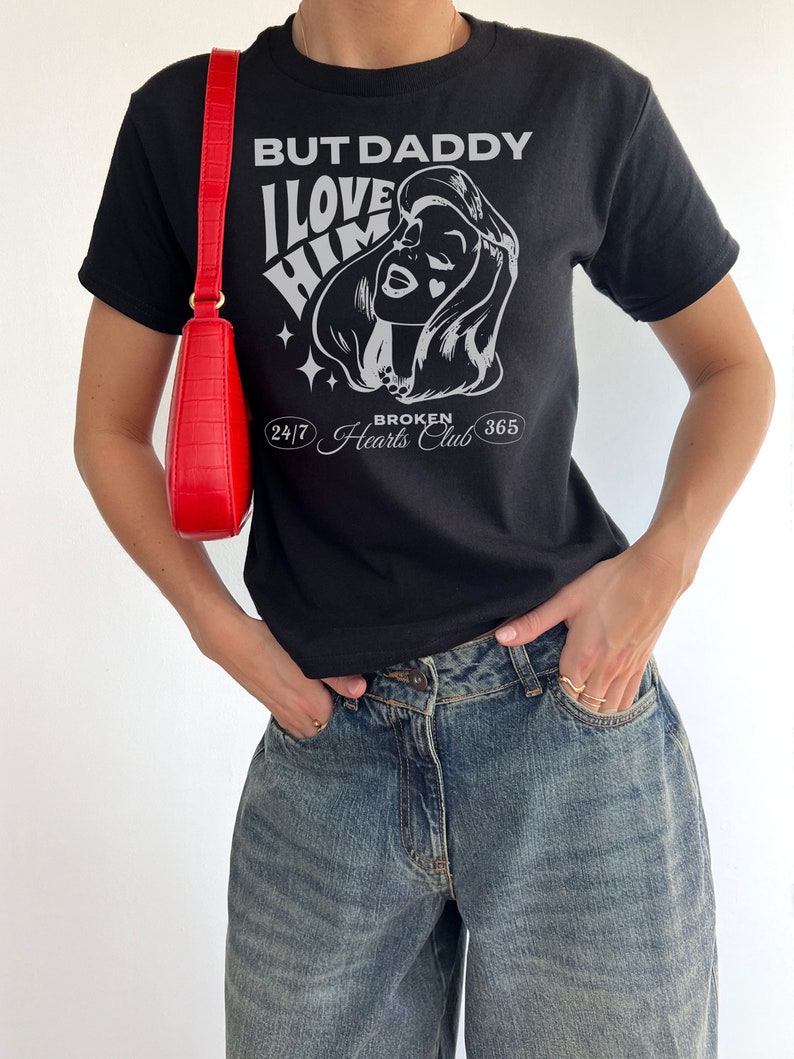 Puede incluir: Camiseta negra con un estampado gr&aacute;fico de la cara de una mujer y el texto "But Daddy I Love Him 24/7 Broken Hearts Club 365".