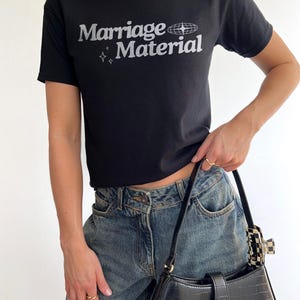 Puede incluir: Una camiseta negra con el texto "Marriage Material" en blanco y un peque&ntilde;o gr&aacute;fico de un globo terr&aacute;queo con estrellas alrededor.