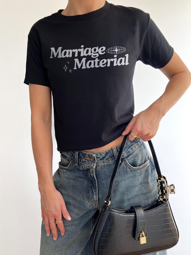Puede incluir: Una camiseta negra con el texto "Marriage Material" en blanco y un peque&ntilde;o gr&aacute;fico de un globo terr&aacute;queo con estrellas alrededor.