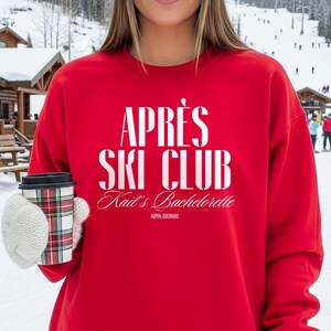 Op de afbeelding: Een rode sweatshirt met de witte tekst "APRÈS SKI CLUB". Daaronder staat "Kait's Bachelorette" en "ASPEN, COLORADO". De persoon houdt een koffiekopje met een ruitpatroon vast en draagt witte wanten.