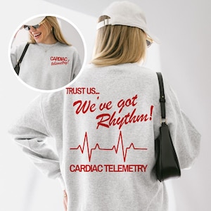 Peut inclure: Un sweat-shirt gris clair avec du texte rouge et un graphique de battement de cœur. Le devant du sweat-shirt indique "CARDIAC telemetry!" et le dos indique "TRUST US... We've got Rhythm!" avec une ligne de battement de cœur rouge et "CARDIAC TELEMETRY".