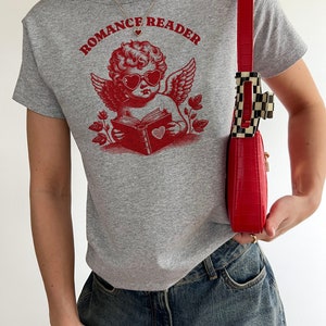 May include: A grey t-shirt with a red graphic of a cherub wearing heart-shaped sunglasses and reading a book with a heart on the cover. The text "Romance Reader" is printed above the cherub.