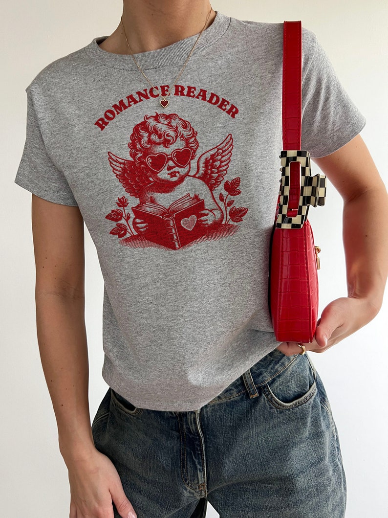 May include: A grey t-shirt with a red graphic of a cherub wearing heart-shaped sunglasses and reading a book with a heart on the cover. The text "Romance Reader" is printed above the cherub.