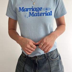 Puede incluir: Una camiseta azul claro con el texto "Marriage Material" en letras azules. El texto est&aacute; acompa&ntilde;ado por un peque&ntilde;o gr&aacute;fico de un globo terr&aacute;queo con estrellas.