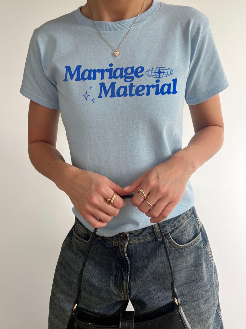 Puede incluir: Una camiseta azul claro con el texto "Marriage Material" en letras azules. El texto est&aacute; acompa&ntilde;ado por un peque&ntilde;o gr&aacute;fico de un globo terr&aacute;queo con estrellas.