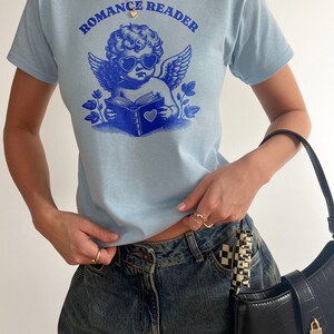 May include: A light blue t-shirt with a graphic of a cherub wearing heart-shaped sunglasses and reading a book. The text "Romance Reader" is printed above the cherub.