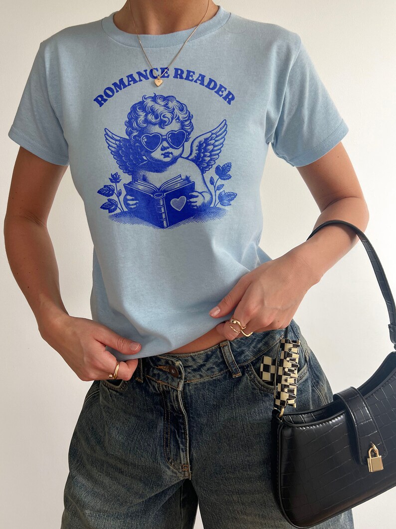 May include: A light blue t-shirt with a graphic of a cherub wearing heart-shaped sunglasses and reading a book. The text "Romance Reader" is printed above the cherub.